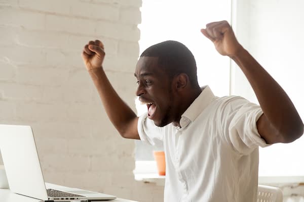 Excited neurodivergent Black man raising his arms while looking at a laptop